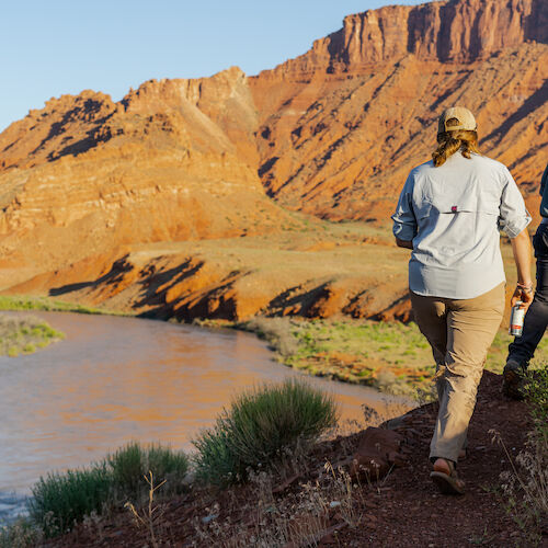 Two people are walking by a river in a desert landscape with red rock formations and clear blue sky.