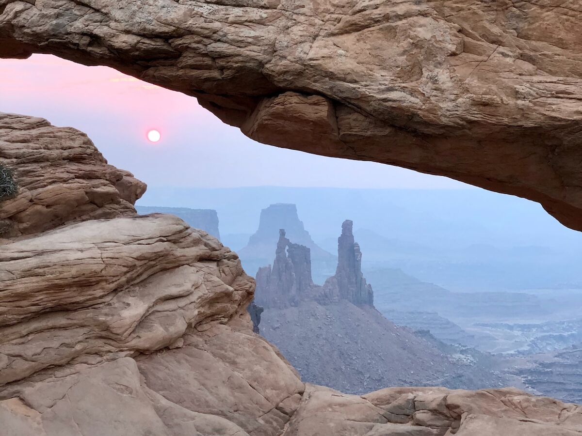 A natural rock arch frames a scenic view of distant rock formations under a pastel sky.