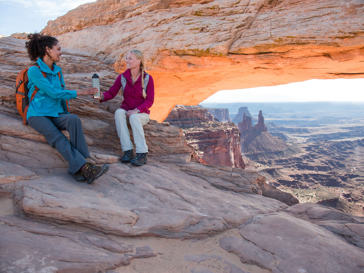 Two people sitting on rocks under a natural arch in a desert landscape, sharing a drink.