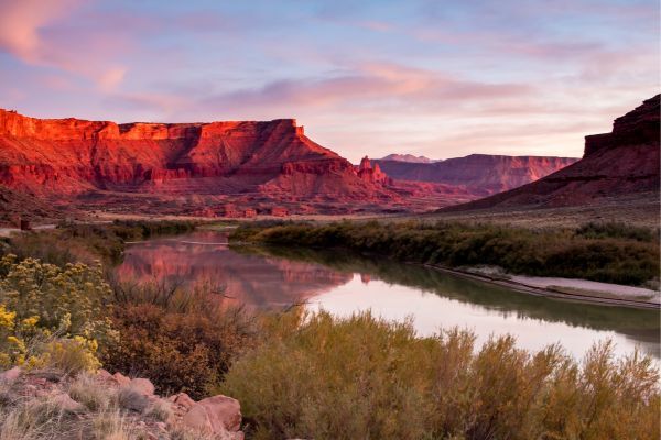 A serene landscape with a winding river reflecting the sky, flanked by desert vegetation and majestic red sandstone cliffs at sunset.