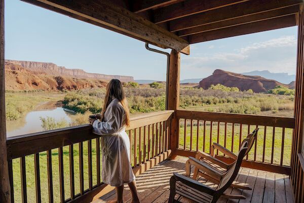 A person in a robe stands on a wooden deck, overlooking a river and red rock landscape under a clear sky.