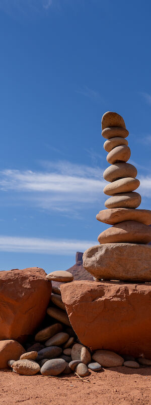 The image shows a stack of smooth stones balanced on a large rock in a desert landscape under a clear blue sky.