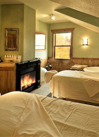 The image shows a cozy spa room with massage tables, a bathtub, a fireplace, and robes hanging on the wall, creating a relaxing ambiance.