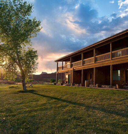 A rustic two-story wooden building with a porch sits beside a tree at sunset, under a sky with scattered clouds and sun rays peeking through.