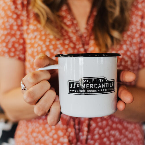 A person holds a white mug with "Mile 17 Mercantile" printed on it, wearing a red patterned shirt.