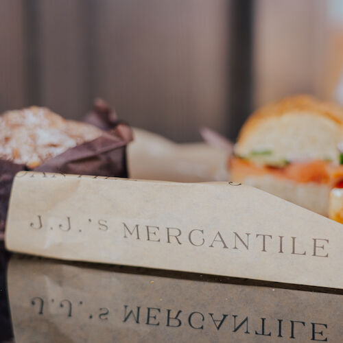 A wrapped sandwich with "J.J.’s Mercantile" branding next to a muffin and a drink on a reflective surface.