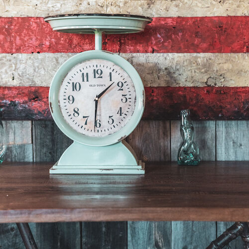 A vintage clock scale on a wooden surface with a rustic striped background, resembling a weathered flag, and a small glass bottle.