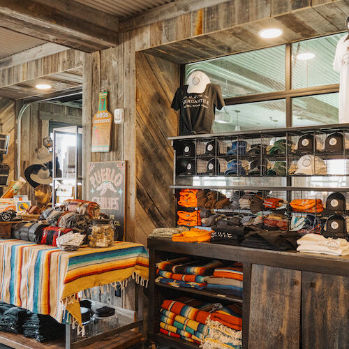 The image shows a rustic store interior with clothing, caps, blankets, and souvenirs displayed on wooden shelves and tables.