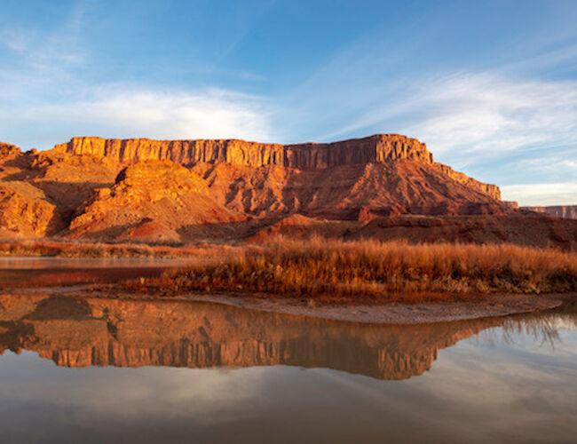 A stunning landscape featuring red rock formations and cliffs, with a calm river below reflecting the scene under a partly cloudy sky.
