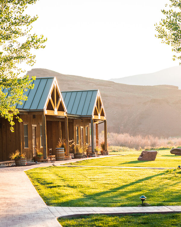 A scenic view of a row of cabins with metal roofs, a lush green lawn, trees, and mountains in the background, bathed in warm sunlight.
