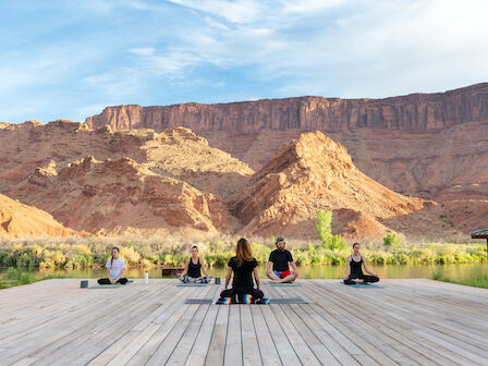 People practice yoga on a wooden deck with red cliffs in the background under a blue sky.