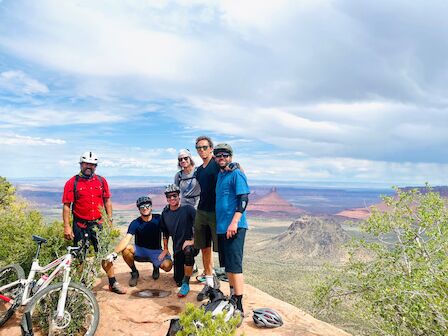 A group of five people in biking gear pose on a scenic viewpoint with mountains and vast landscape in the background.
