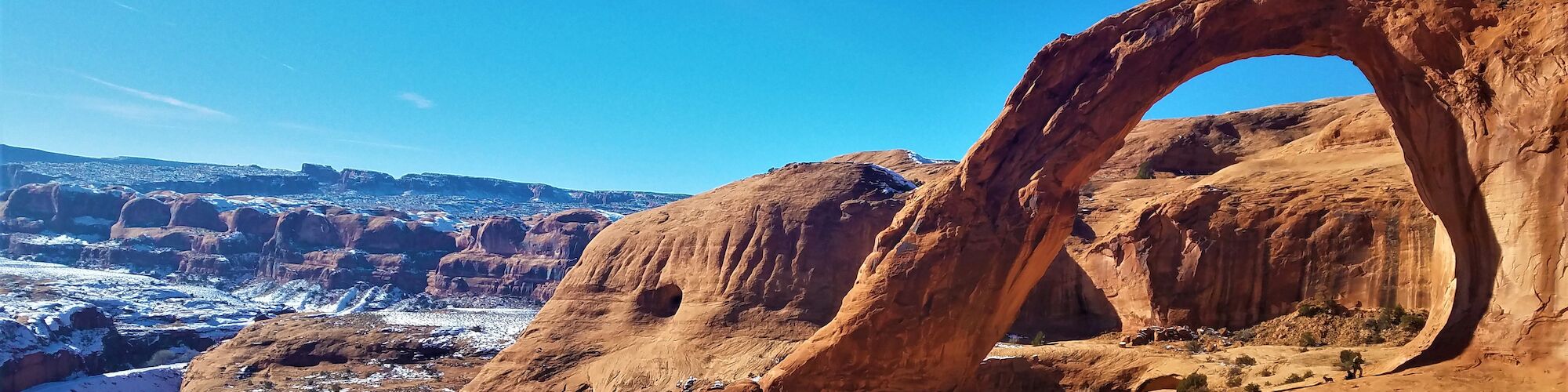 A natural sandstone arch stands prominently against a clear blue sky in a rocky, desert landscape with distant snow-dusted hills.