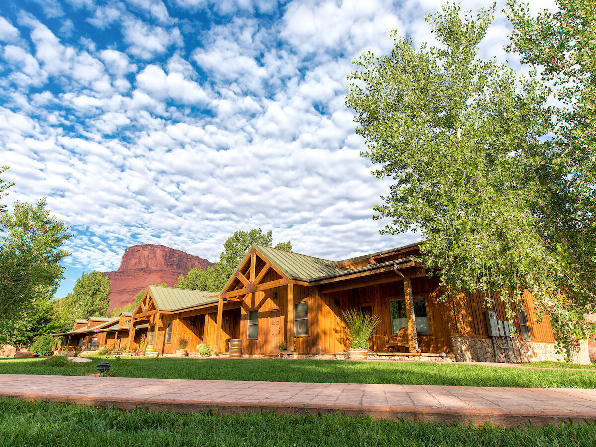 A rustic building with wooden accents is surrounded by trees and grass, under a sky with scattered clouds and distant red rock formations.