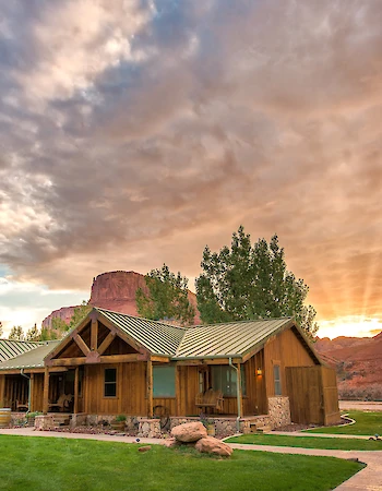 A rustic building with a green roof set against a dramatic sunset sky, surrounded by trees and a manicured lawn with a paved path.