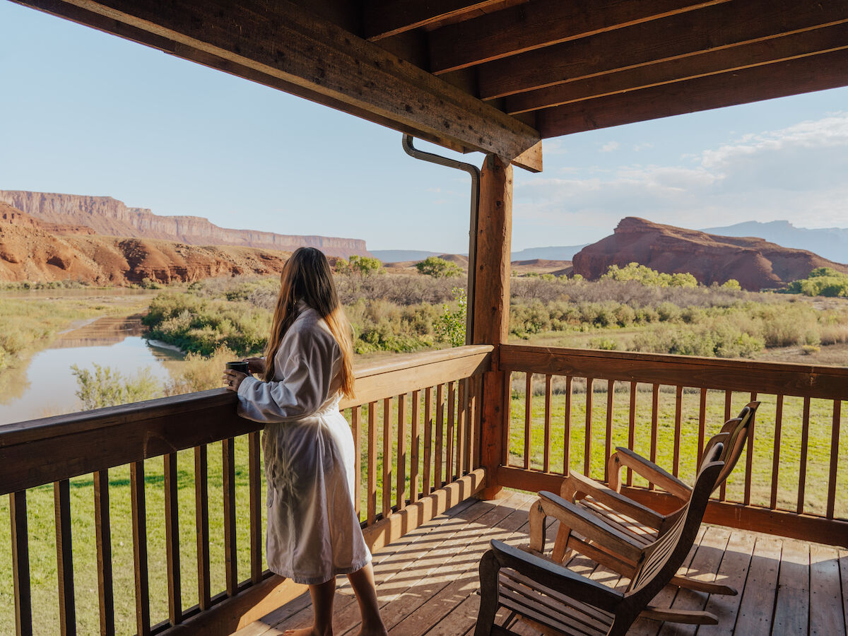 A person in a robe stands on a wooden deck, looking at a scenic view of a river and red rock formations under a clear sky.
