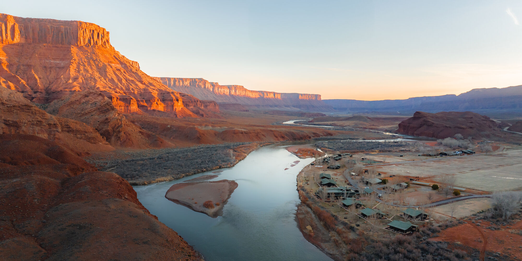 A river winds through a desert landscape with red cliffs and sparse vegetation, captured during sunset.