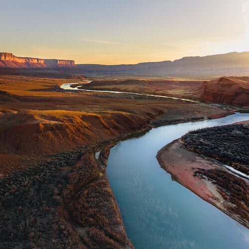 A winding river flows through a canyon landscape at sunset, with distant mountains and a clear sky creating a serene scene.