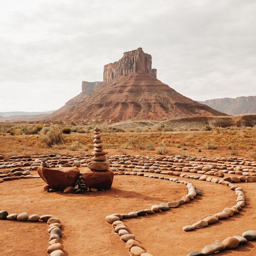 A rocky, desert landscape with a central rock formation surrounded by a stone labyrinth on sandy ground.
