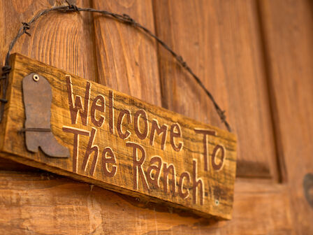 A wooden sign on a door reads, "Welcome to The Ranch" with a boot design, hung by wire.