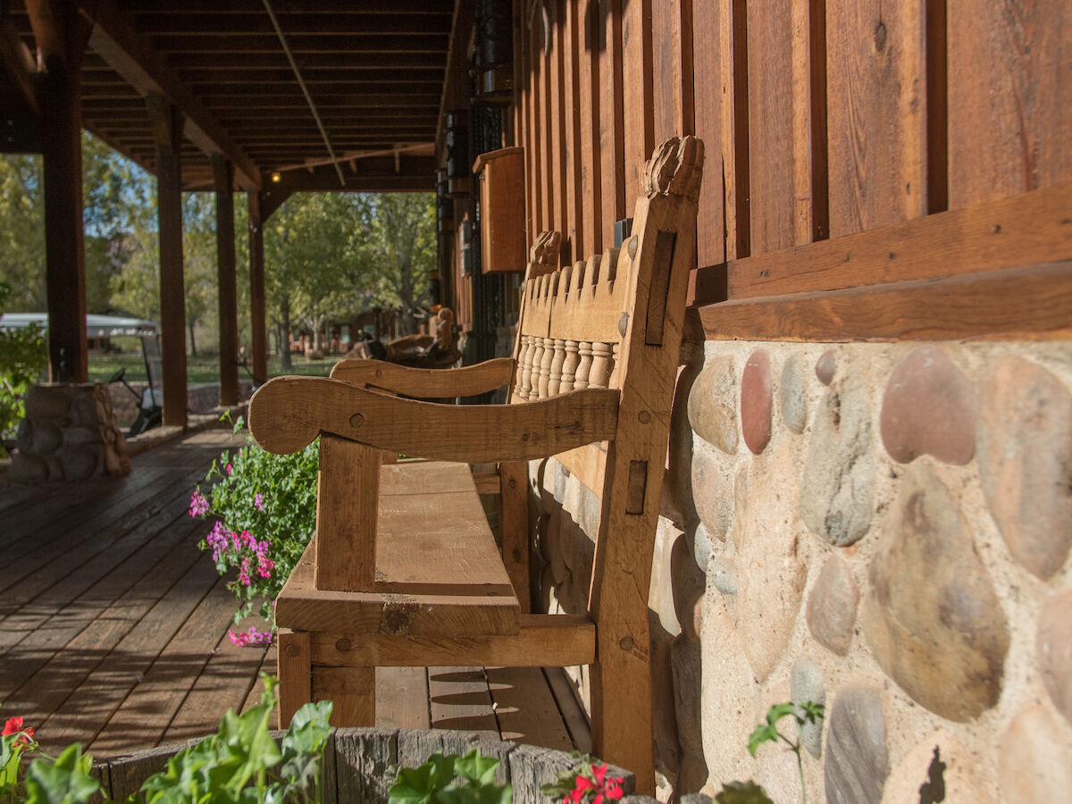 A wooden porch with a long bench along a stone wall, surrounded by plants and greenery under a clear sky on a sunny day.