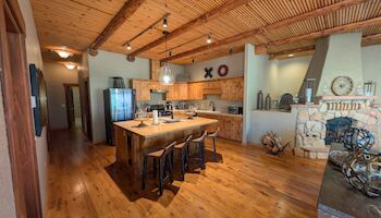 A rustic kitchen with wooden floors and ceiling, a central island, bar stools, and a stone fireplace, creating a cozy atmosphere.