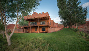 A large two-story adobe house with balconies sits on a grassy hill, surrounded by trees under a clear sky.