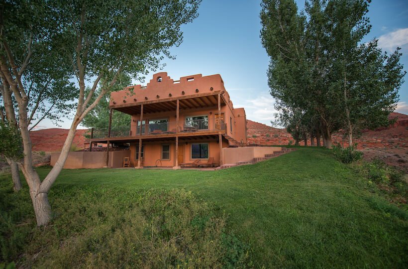 A large two-story adobe house with balconies sits on a grassy hill, surrounded by trees under a clear sky.