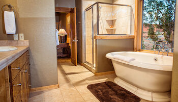 The image shows a bathroom with a bathtub, shower, vanity, and a large window overlooking a view, featuring warm tones and wood accents.