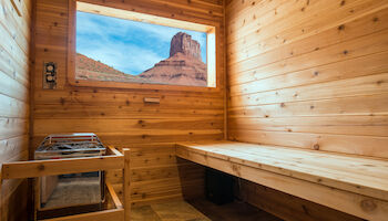A wooden sauna with a bench and heater, featuring a window view of a desert rock formation against a blue sky.