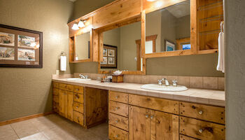 The image shows a bathroom with wooden cabinets, a large mirror, and framed artwork on the wall. There's warm lighting from wall fixtures.