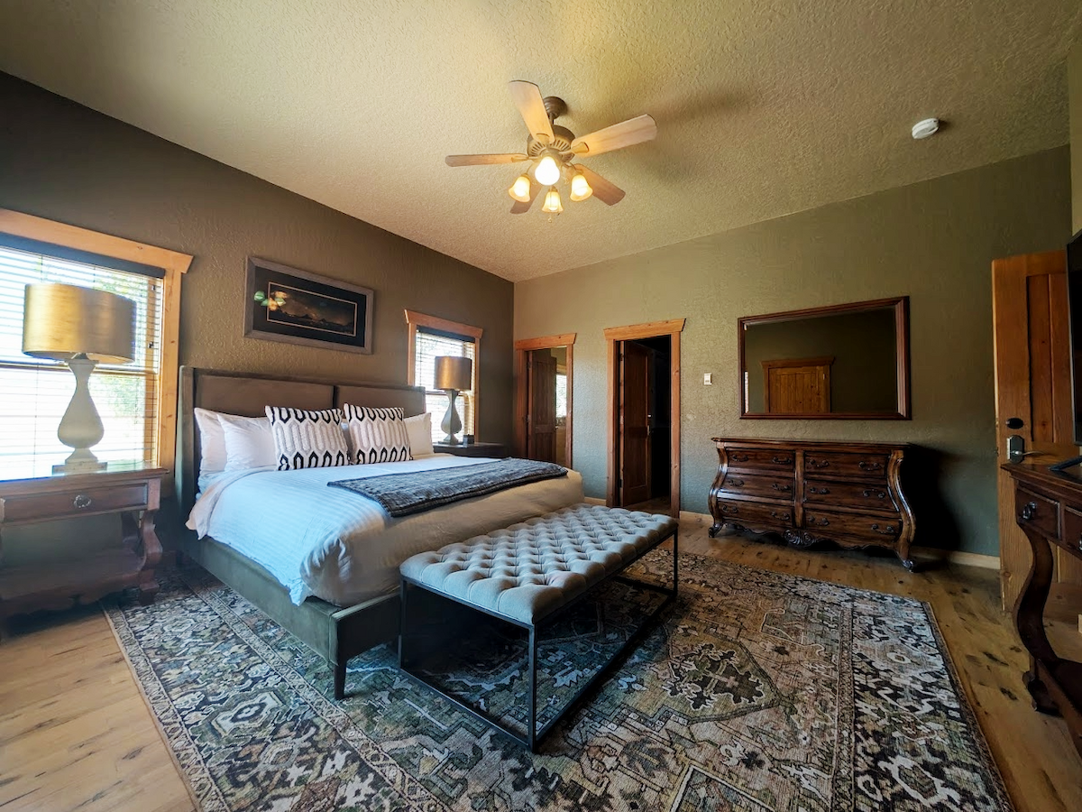 A cozy bedroom with a large bed, cushioned bench, wooden dressers, windows, elegant lamps, and a patterned rug.