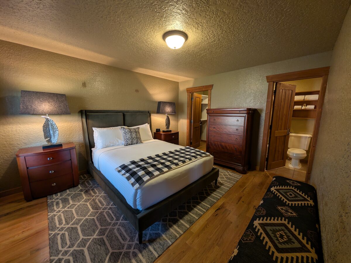 A cozy bedroom with a neatly made bed, wooden furniture, decorative lamps, and a patterned rug. A bathroom is visible through a doorway.