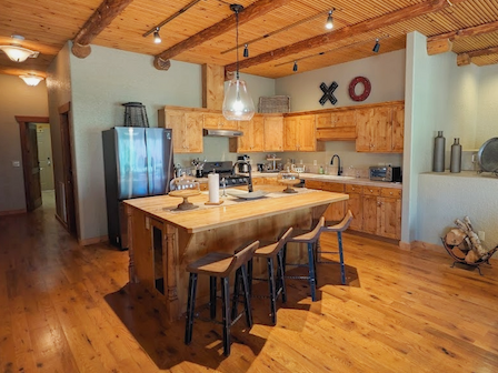 A rustic kitchen with wooden cabinets, a central island, stools, and stainless steel appliances under a wood-beamed ceiling.