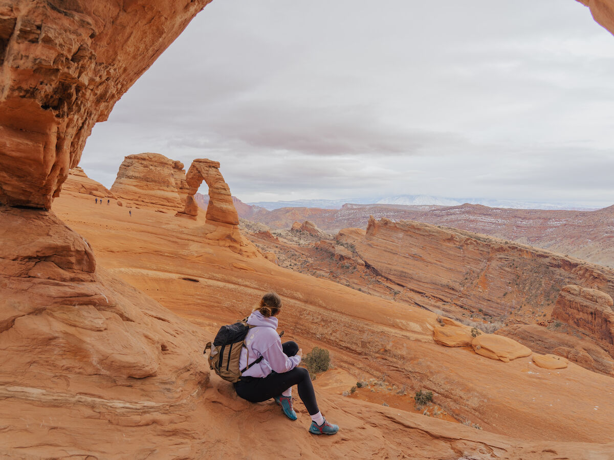A person sits on a rocky ledge, gazing at the Arches National Park landscape with Delicate Arch in the distance on a cloudy day.
