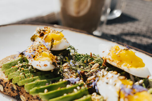 Avocado toast topped with sliced eggs and seeds on a plate, accompanied by a frothy coffee in a glass mug in the background.