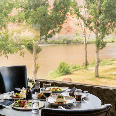 A round dining table with meals and drinks is set near a large window offering a scenic view of trees and a river outside the restaurant.