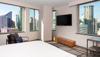 A modern hotel room with a bed, chair, TV, and stunning cityscape views from large windows on multiple sides.
