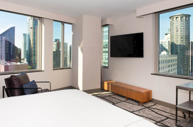 A modern hotel room with a bed, chair, TV, and stunning cityscape views from large windows on multiple sides.