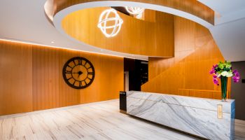 A modern interior with a marble reception desk, wooden walls, a large clock, geometric chandelier, and a vase of flowers on the counter.