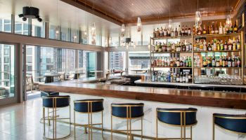 A modern bar has a wooden counter, blue and gold bar stools, shelves with various bottles, pendant lights, and large windows with city views.