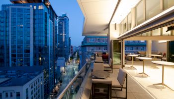 A modern cityscape view from a rooftop terrace at dusk, featuring outdoor seating and tables, with nearby high-rise buildings lit up.