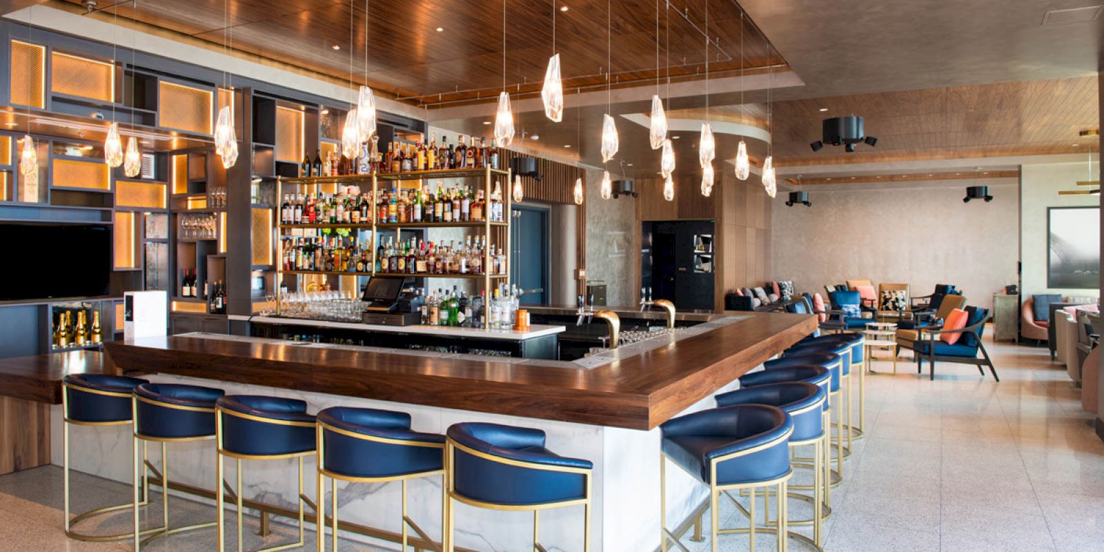 An upscale bar with a modern interior, featuring a wooden bar top, shelves stocked with liquor, blue barstools, and pendant lighting.