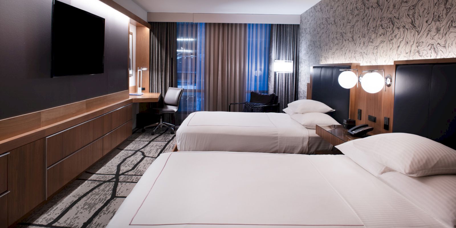 A modern hotel room features two neatly made beds, a wall-mounted TV, a desk with a chair, and contemporary lighting.