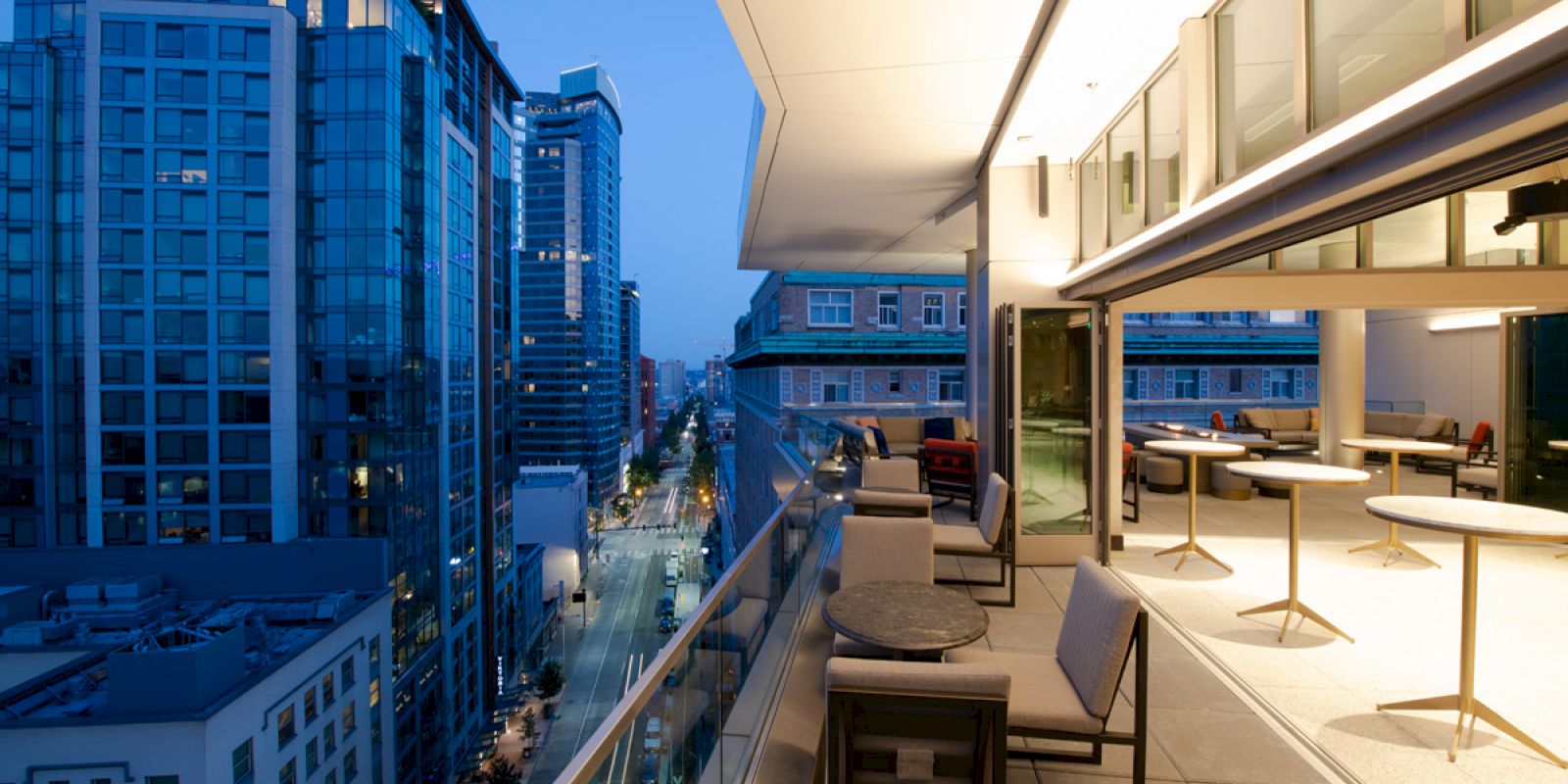 A modern balcony overlooking an urban cityscape at twilight with high-rise buildings, tables, and seating. The scene has a serene, upscale ambiance.