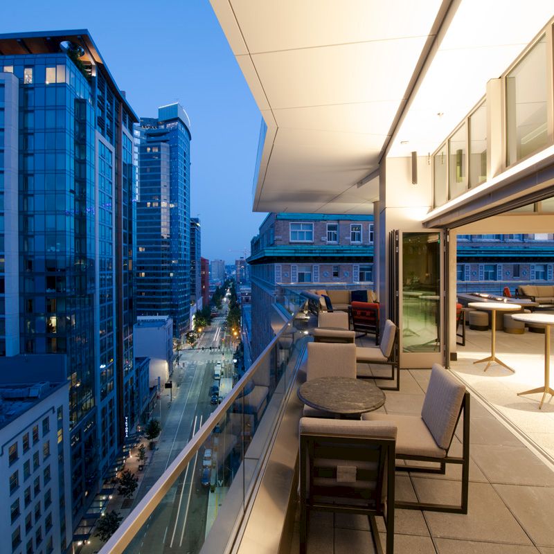 A modern balcony with seating overlooks an urban cityscape featuring tall buildings and streets at twilight, with the interior space brightly lit.