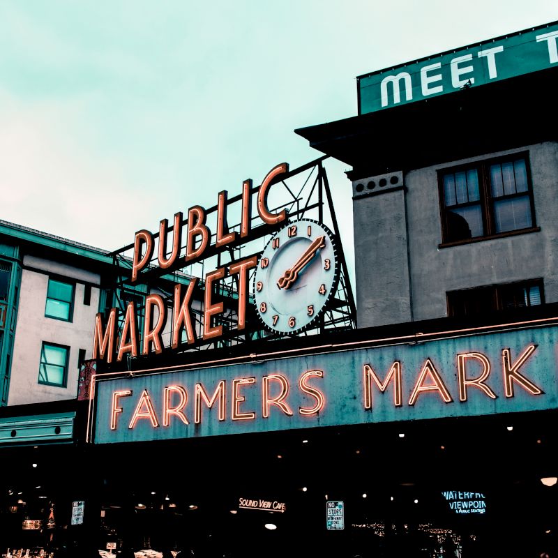 The image shows a city scene with a neon sign for "Public Market" and "Farmers Market," along with a clock, in a commercial area.