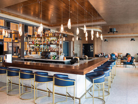 A modern bar with blue cushioned stools, a wooden bar countertop, and hanging pendant lights. The background shows shelves stocked with liquor bottles.