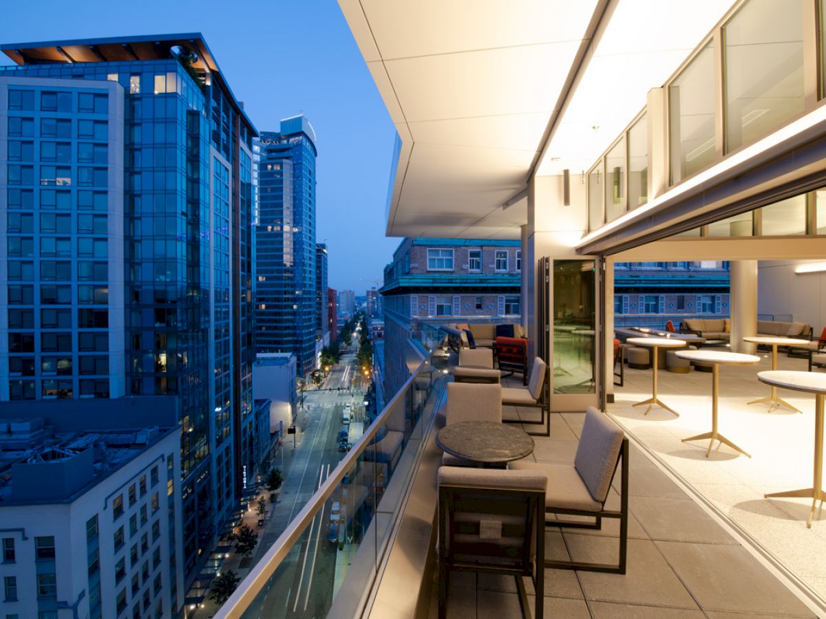 A modern cityscape view showcases high-rise buildings at dusk, with a stylish outdoor balcony featuring seating and tables on the right.