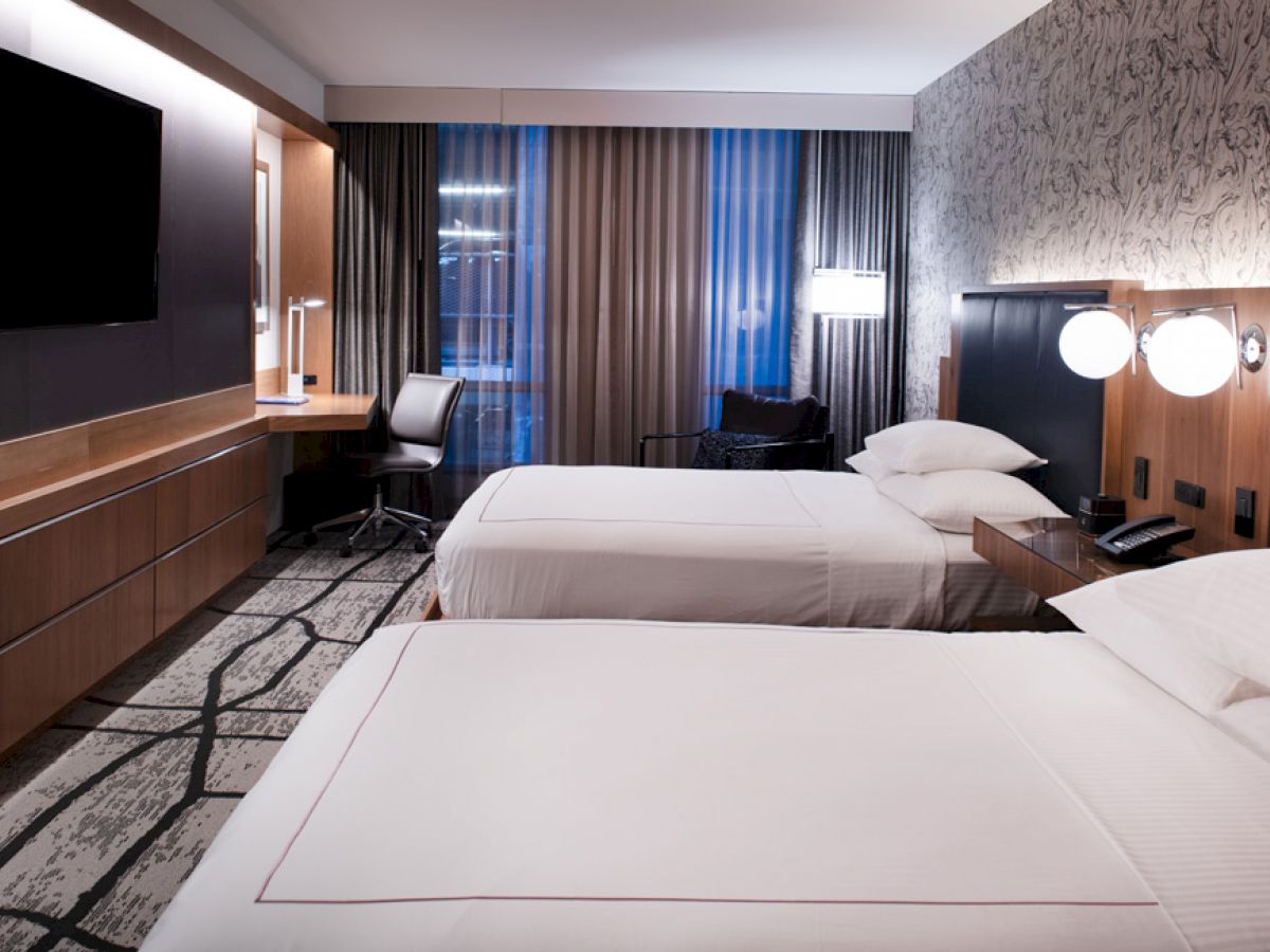 A modern hotel room with two neatly made beds, a television, a work desk, and stylish decor, featuring ambient lighting and large windows.
